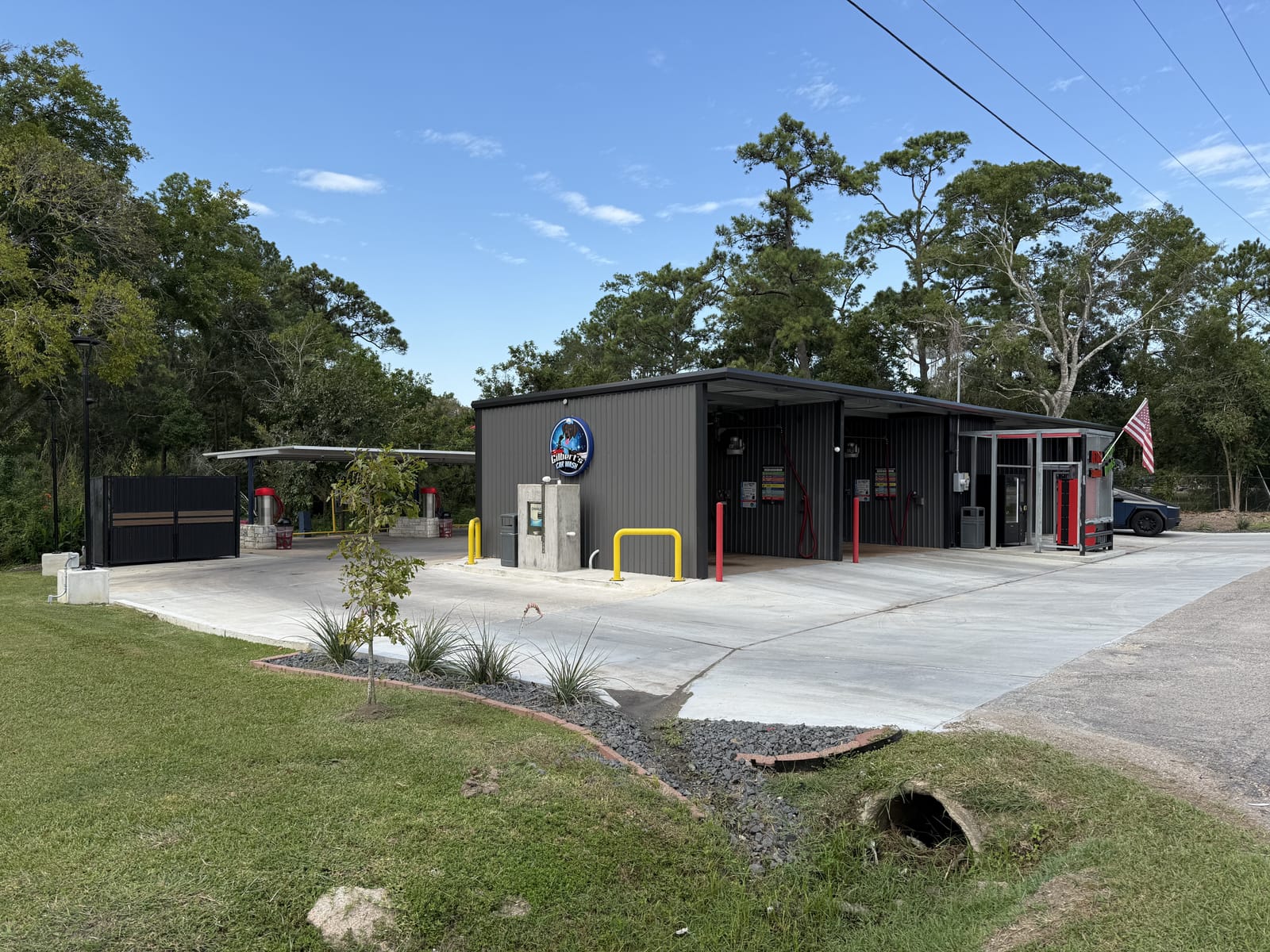 Gilbert's Car Wash in Dickinson, TX — modern dark gray metal building with the Gilbert's logo, self-serve bays, and covered stalls surrounded by trees.